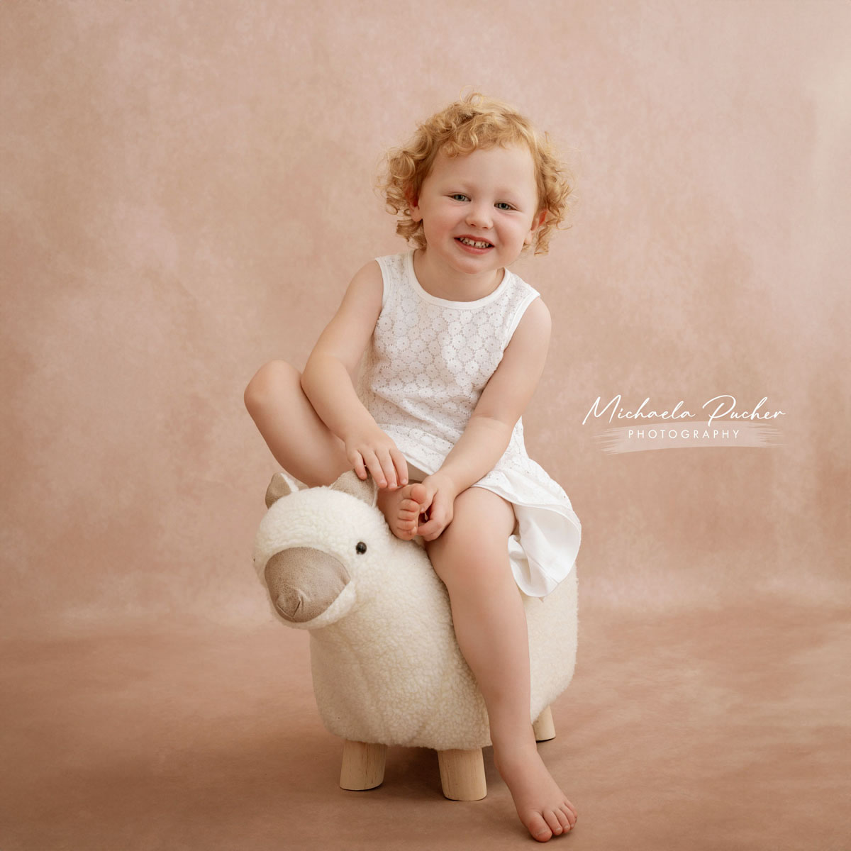 Lachendes blondgelocktes Kind sitzt barfuß auf einem flauschigen Hocker in Tierform vor einem zarten rosigen Hintergrund.